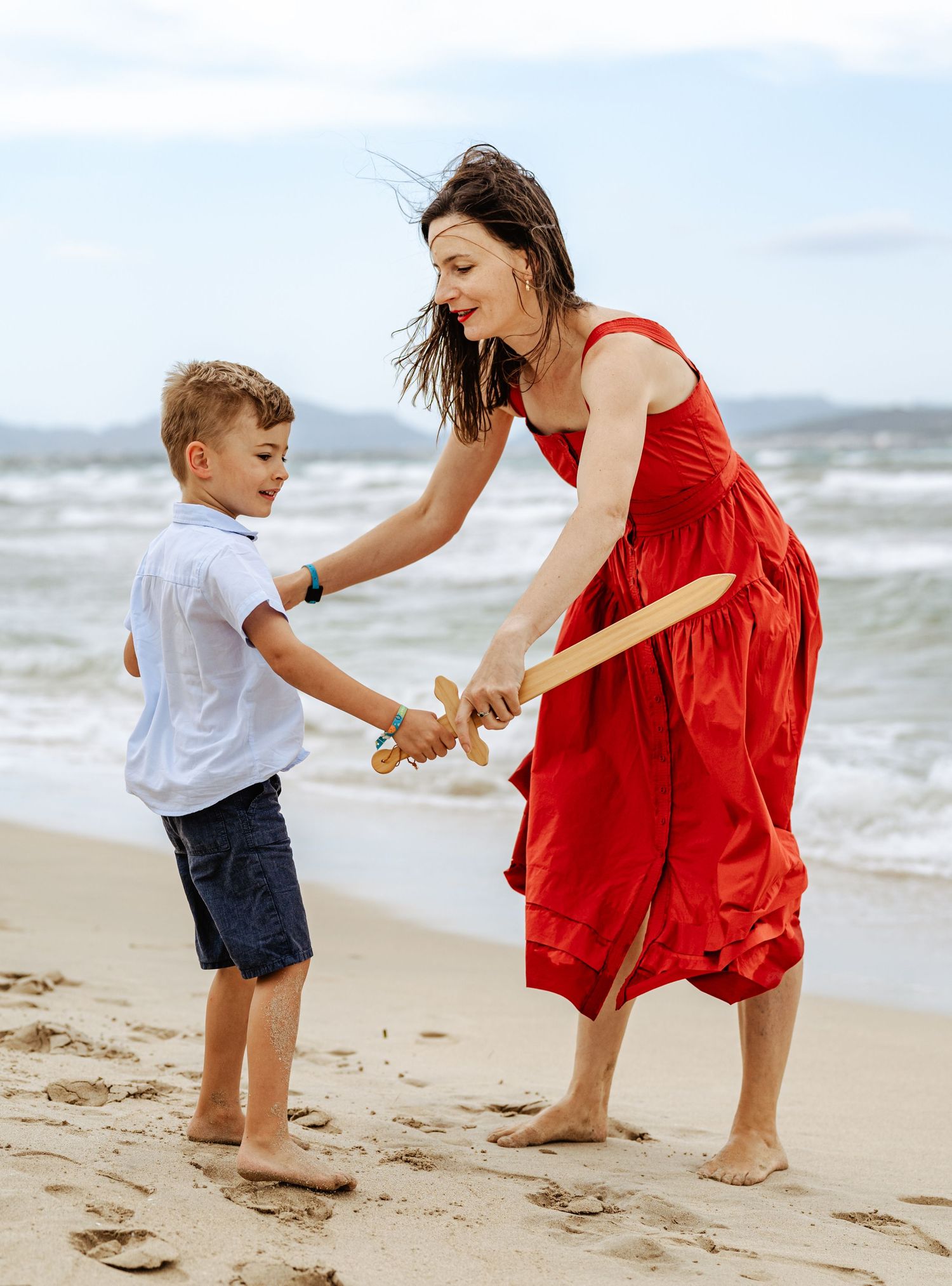 A laughing woman in a red dress and young boy engage in a playful wooden sword fight on the beach.