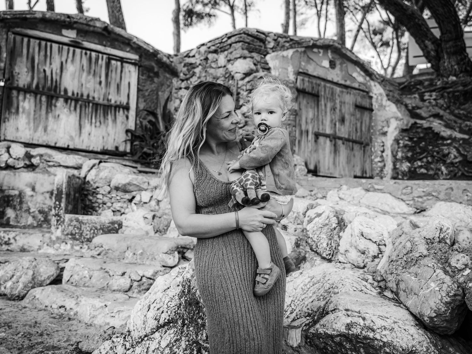 Black and white photo of mother kissing toddler while seated on rocks near rustic stone structures.