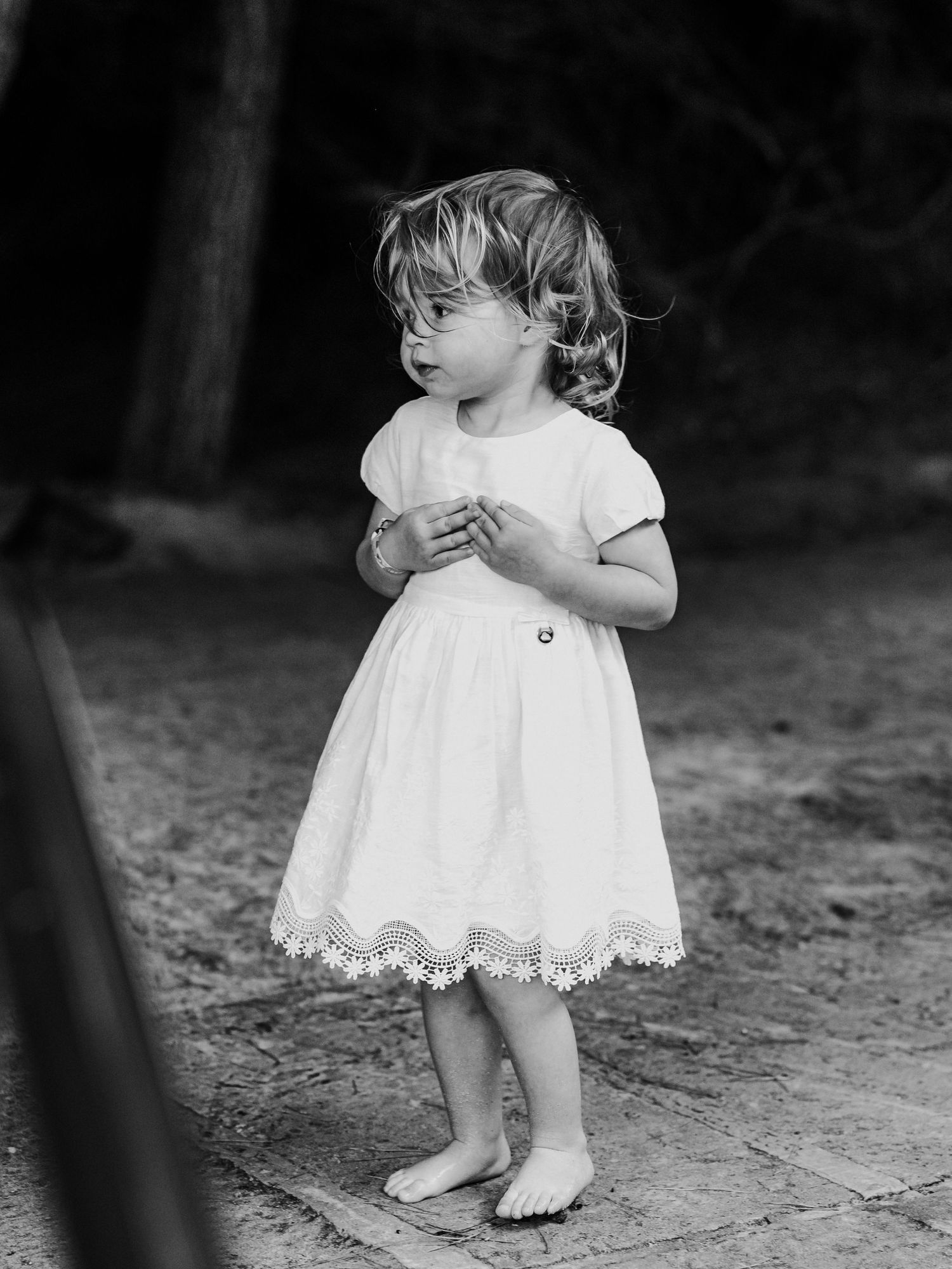 Black and white portrait of toddler girl in white dress standing barefoot outdoors.