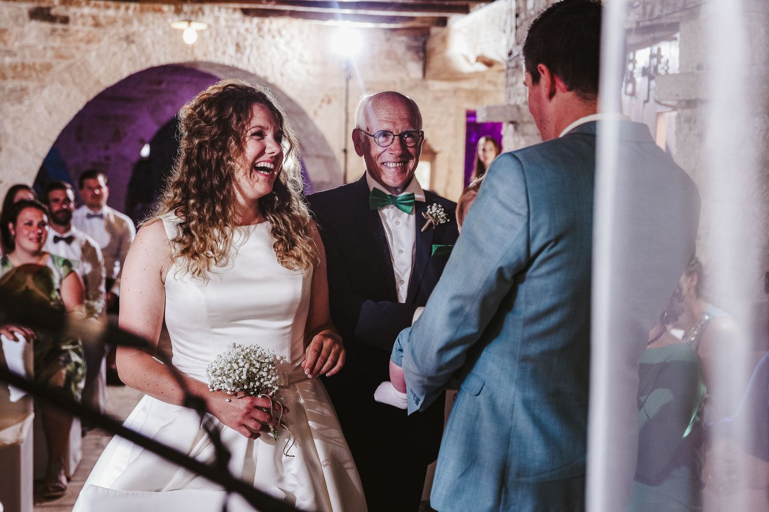 Joyful bride in white dress laughing with her father and groom inside a rustic stone venue.