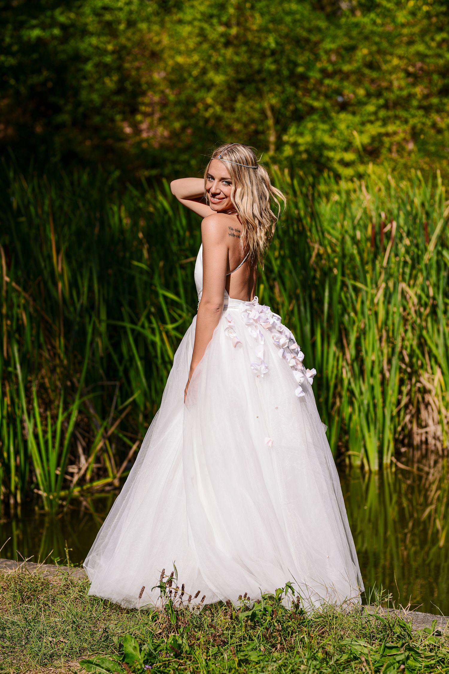 Blonde bride in a backless white floral gown posing gracefully near a serene green pond.