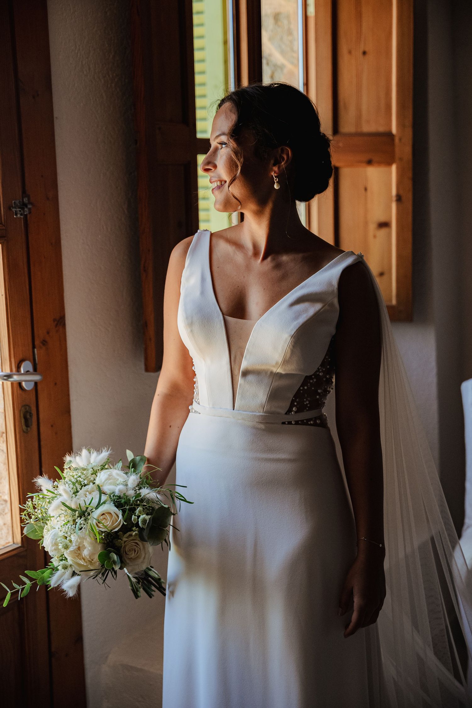 Elegant bride in white gown holding bouquet of white roses gazing out a wooden window.