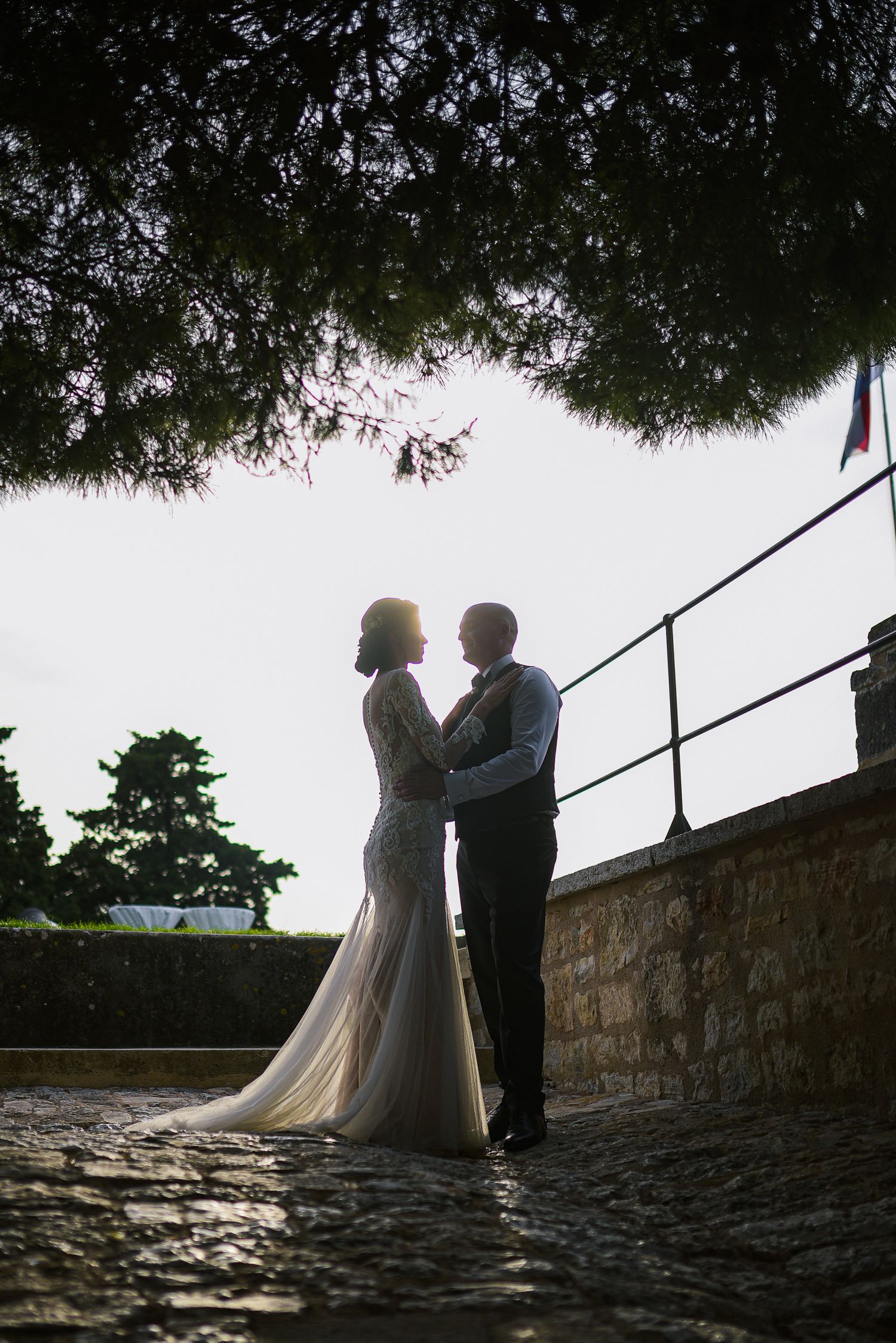 Silhouette of bride and groom embracing on cobblestone path under trees at golden hour.