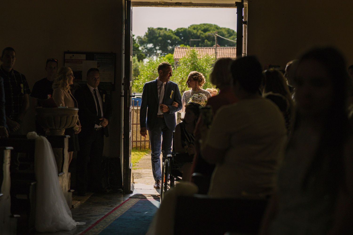 Wedding guests seated inside venue watching bride and groom enter through sunlit doorway.