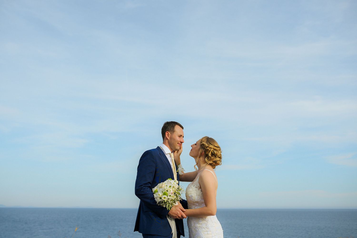 Smiling bride and groom share a joyful moment together with the vast blue ocean and sky behind them.