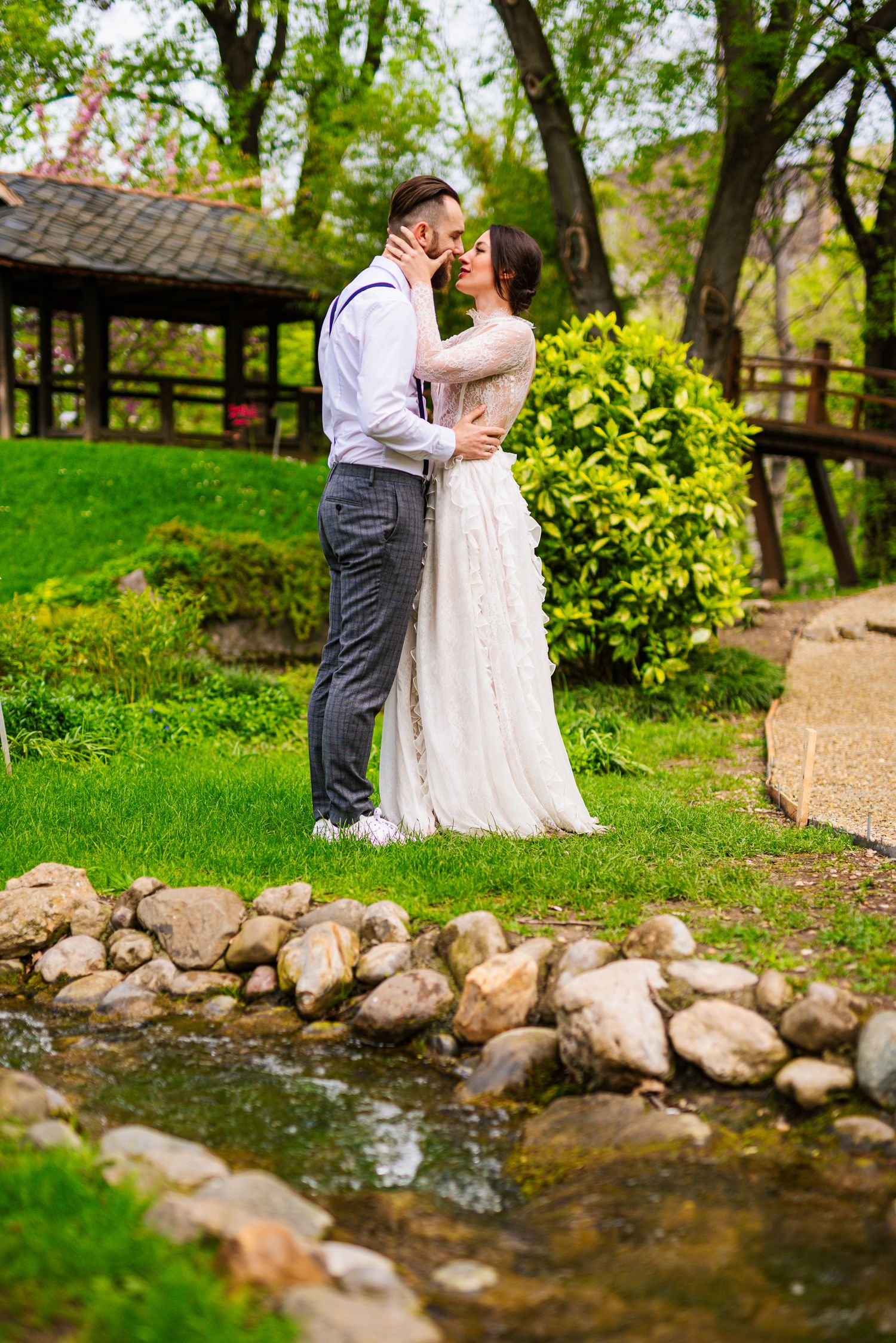 Bride and groom sharing a kiss beside a small rocky stream in a lush green park garden.