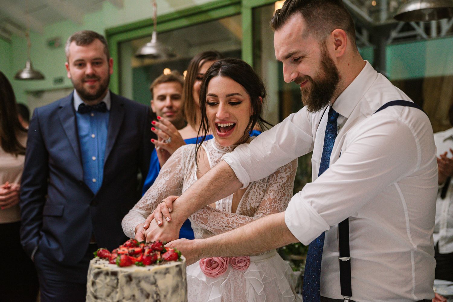Bride and groom cutting strawberry wedding cake together while guests watch and celebrate.