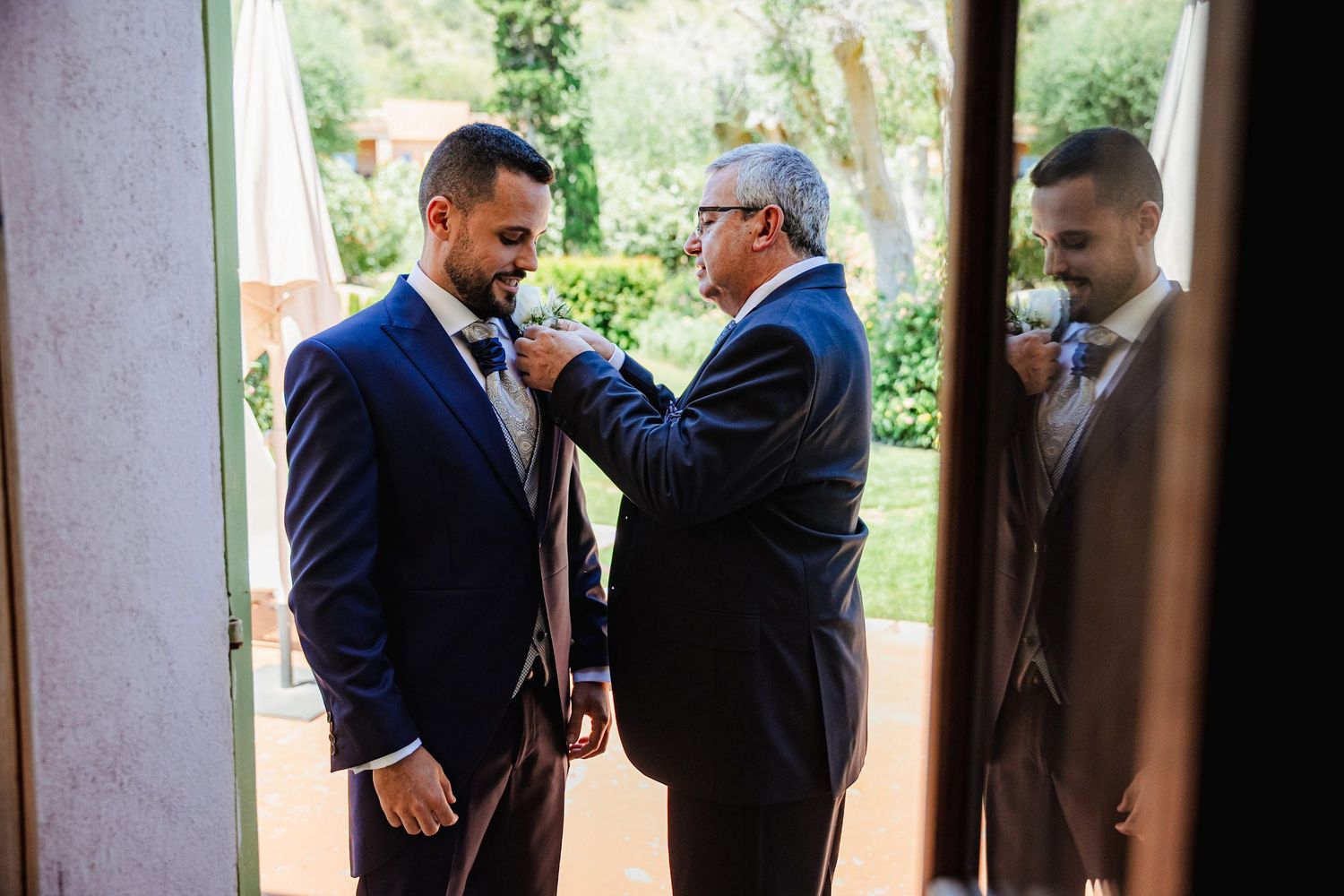 Groom in navy suit having tie adjusted by older man near garden doorway.