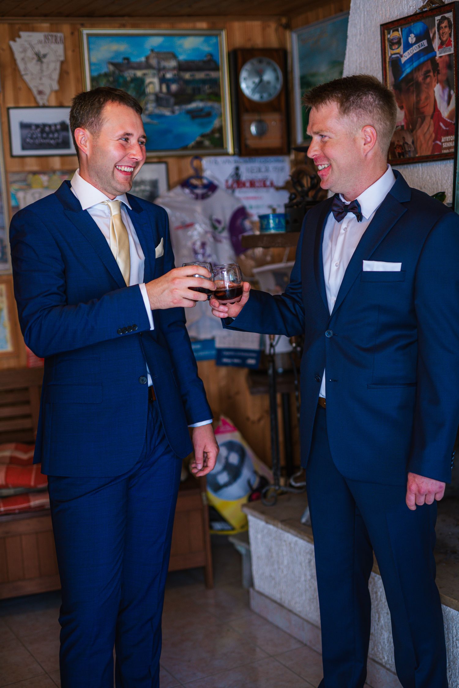 Two men in navy suits toast with drinks while laughing in a decorated room.