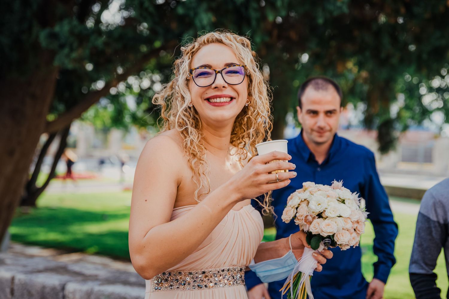 Smiling curly-haired bride in blush gown holding white bouquet outdoors at sunny wedding celebration.