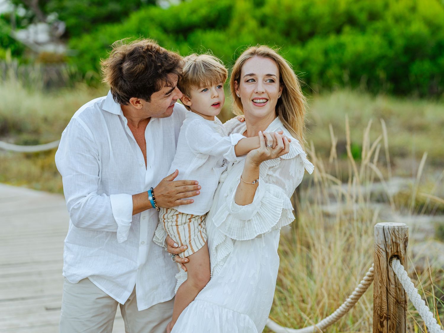 Joyful family of three laughing together outdoors on a wooden boardwalk in white outfits.