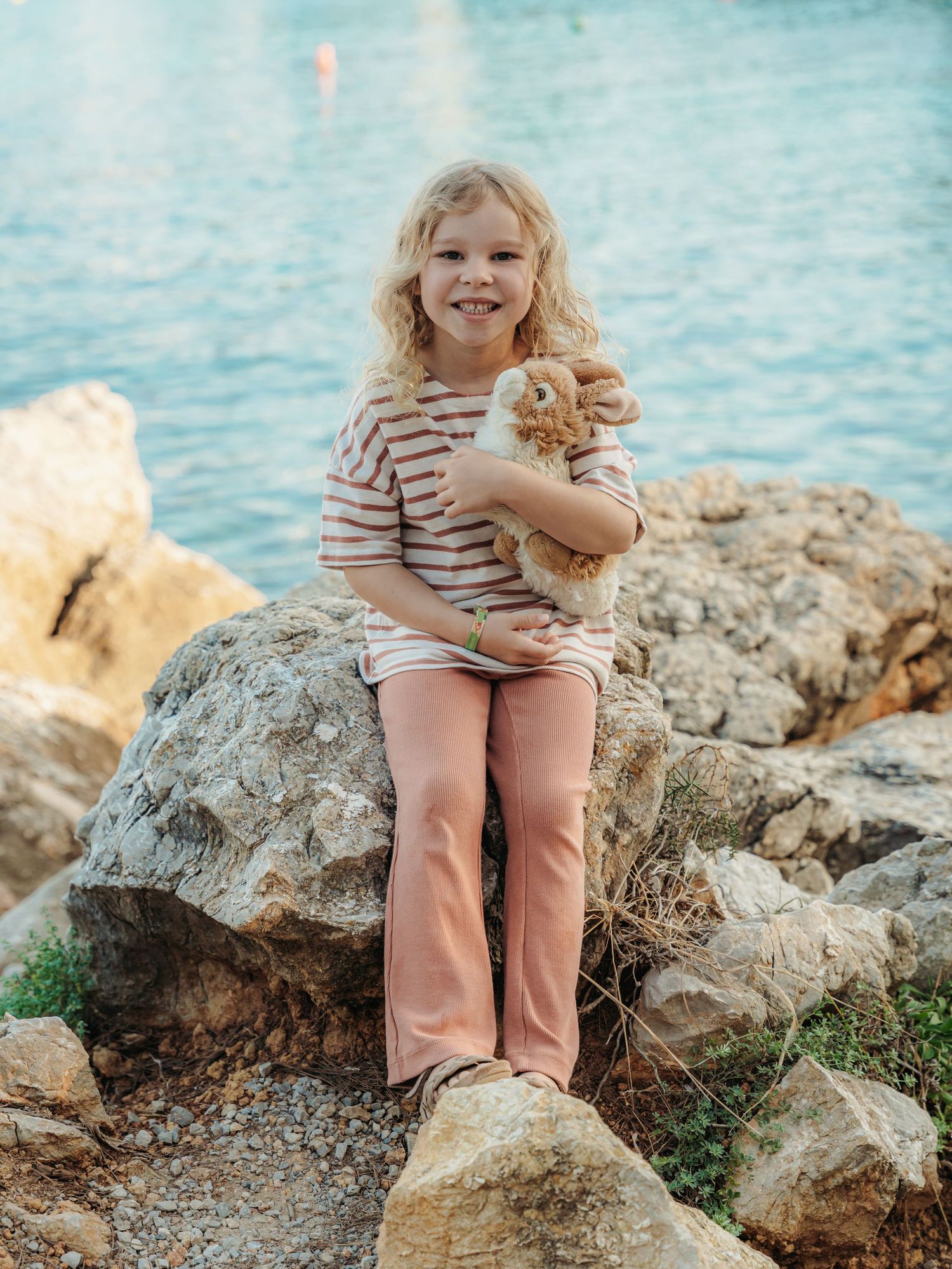 Happy blonde girl in striped top holding stuffed rabbit toy while sitting on rocky shore.