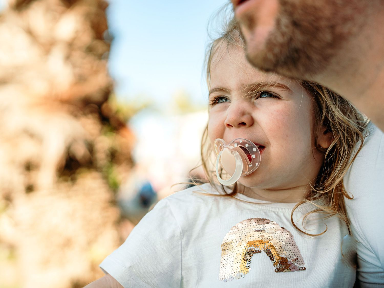Close-up of a toddler girl with a pacifier wearing a sequin top being held by her father outdoors.