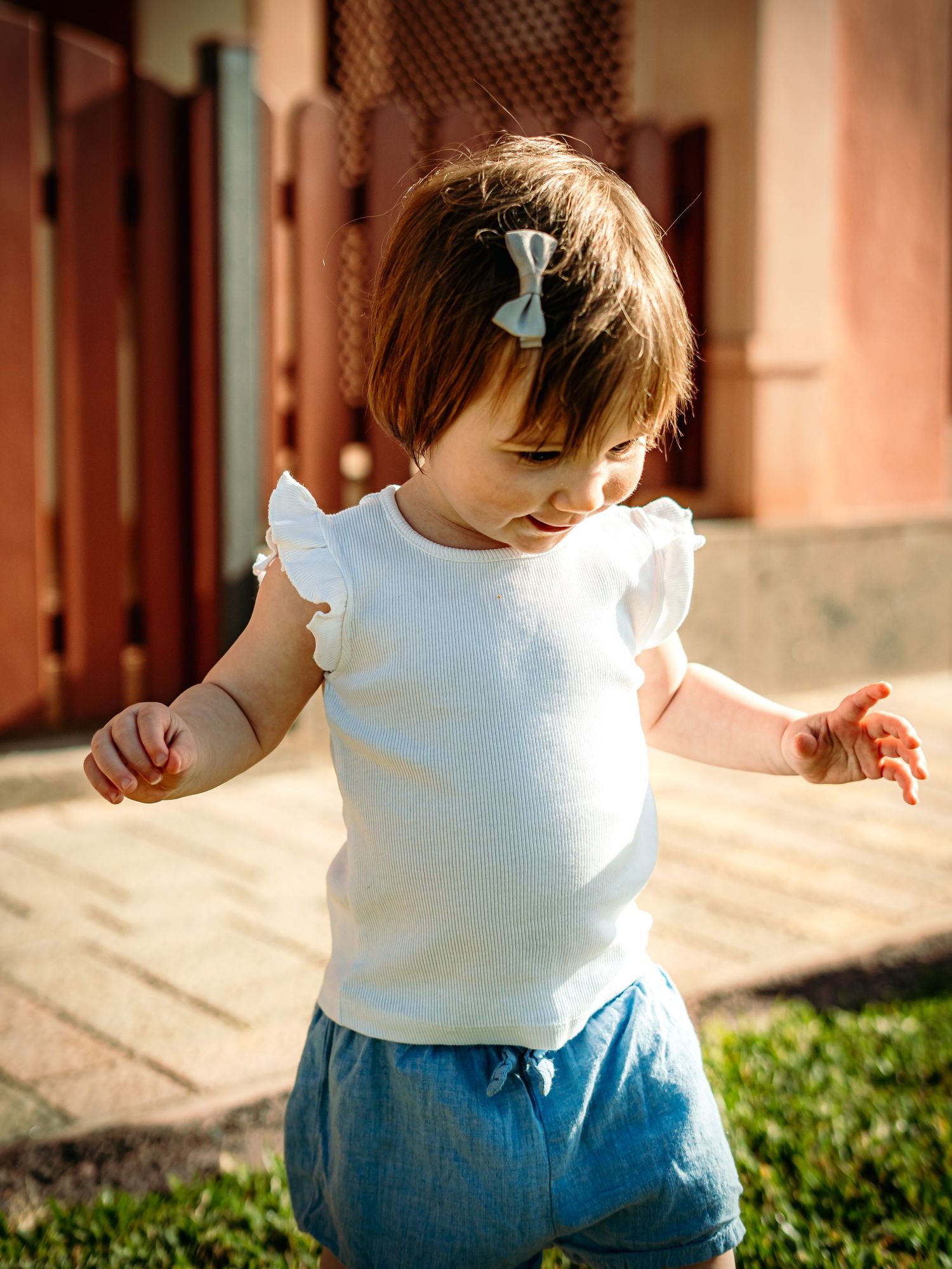 Sweet baby girl in white ruffled top and denim shorts with a bow clip walking outdoors in sunlight.