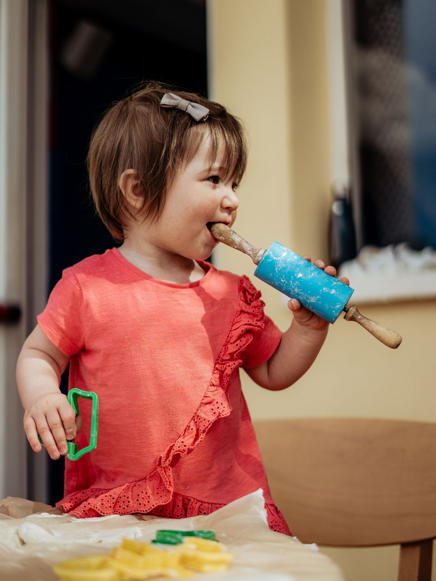Adorable toddler girl in coral dress licking a blue rolling pin while playing with cookie dough.
