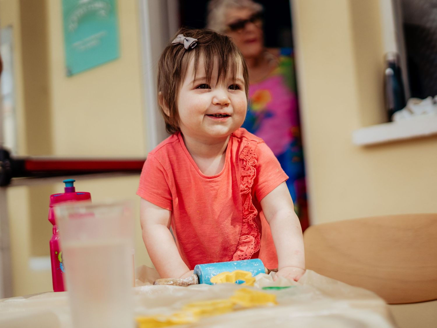 Happy toddler girl in coral shirt sitting at a craft table smiling with supplies around her.