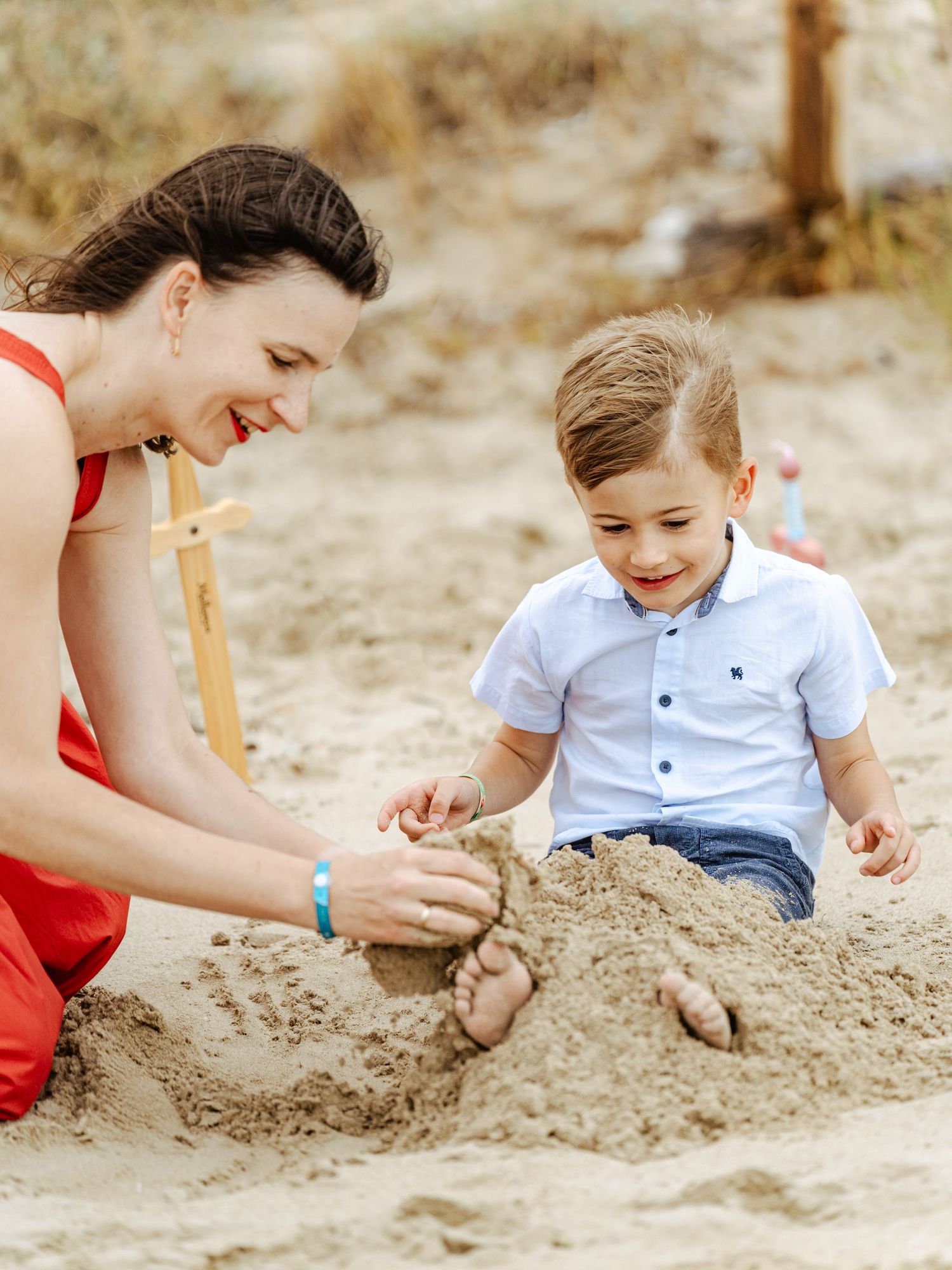 Smiling mother in red dress and little boy playing in sand at the beach together.
