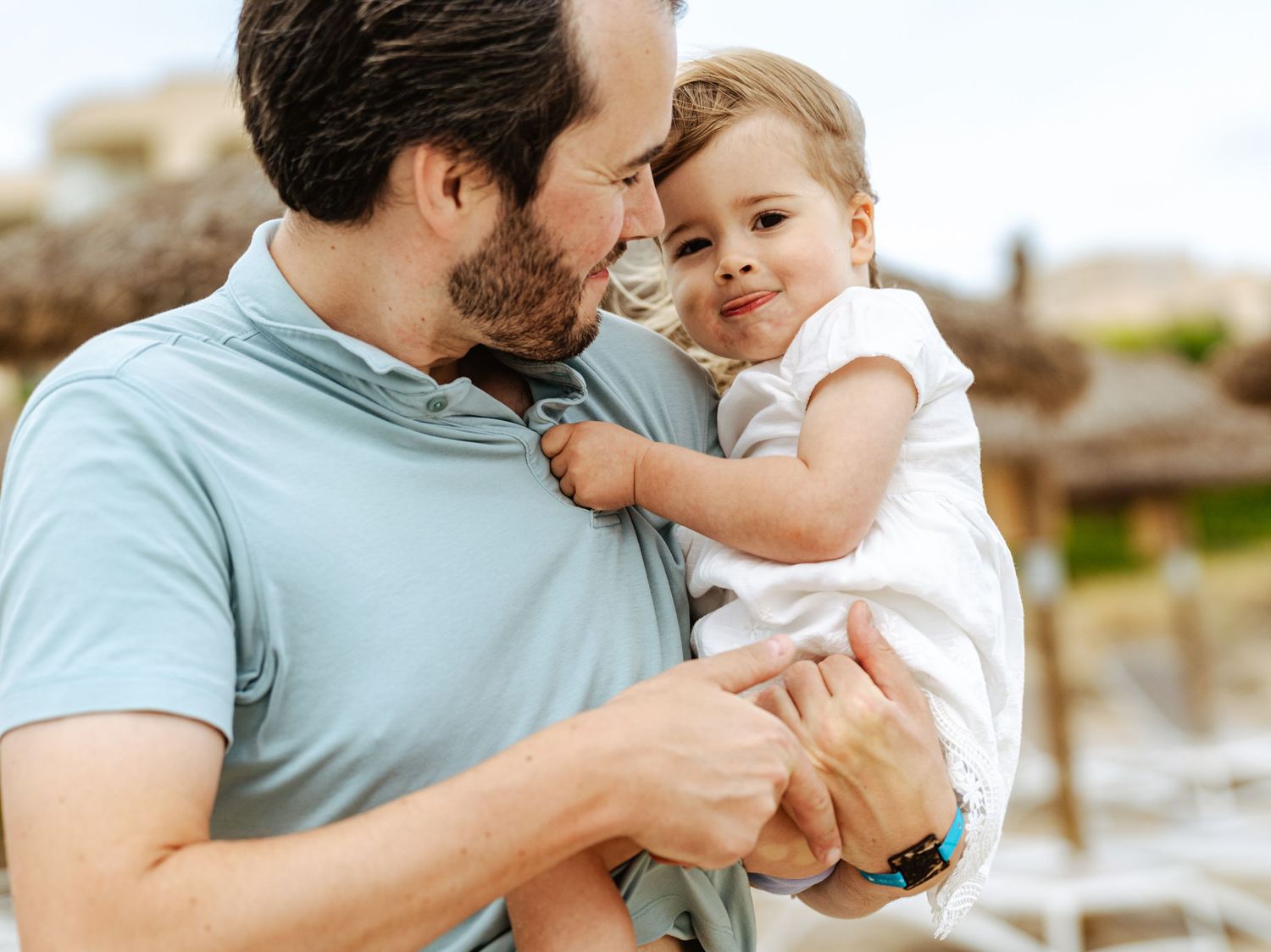 A loving father in a blue shirt holds his happy toddler daughter in a white dress outdoors.
