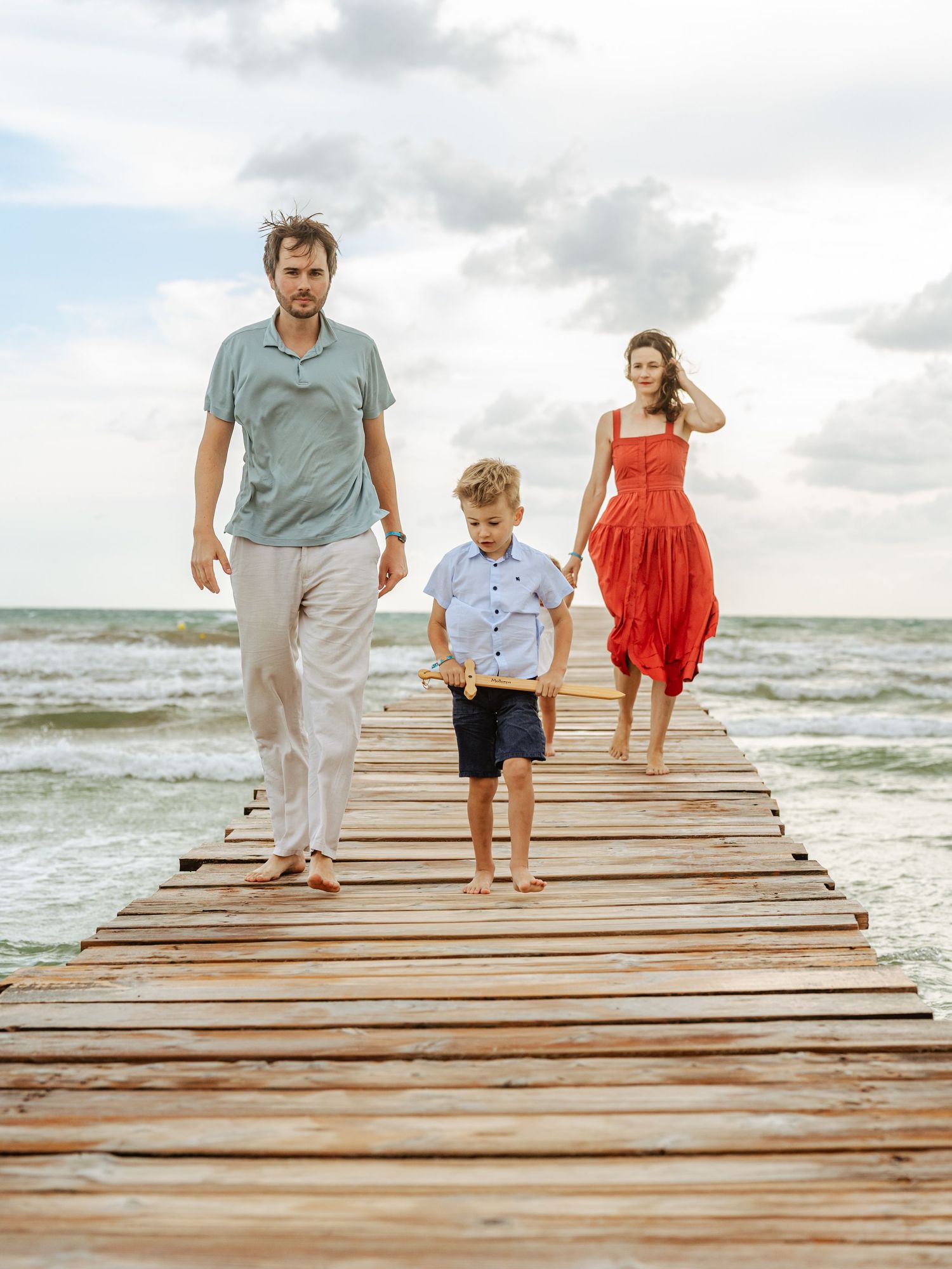 Family of three walking on wooden pier at beach, father and son in front, mother in red dress behind.