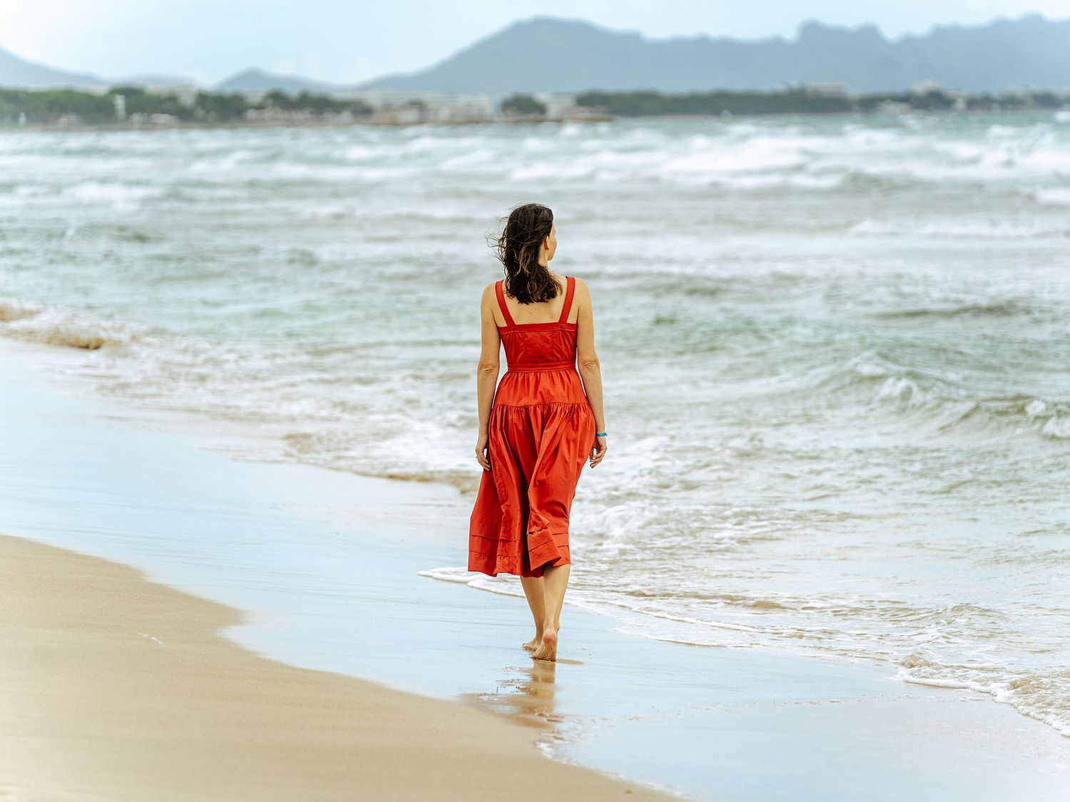 Woman in red midi dress walking barefoot along ocean shoreline on sandy beach.