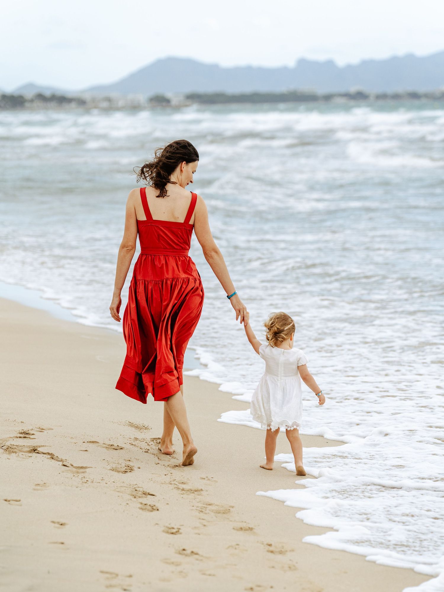 Mother in red dress and toddler girl walk hand in hand along beach.