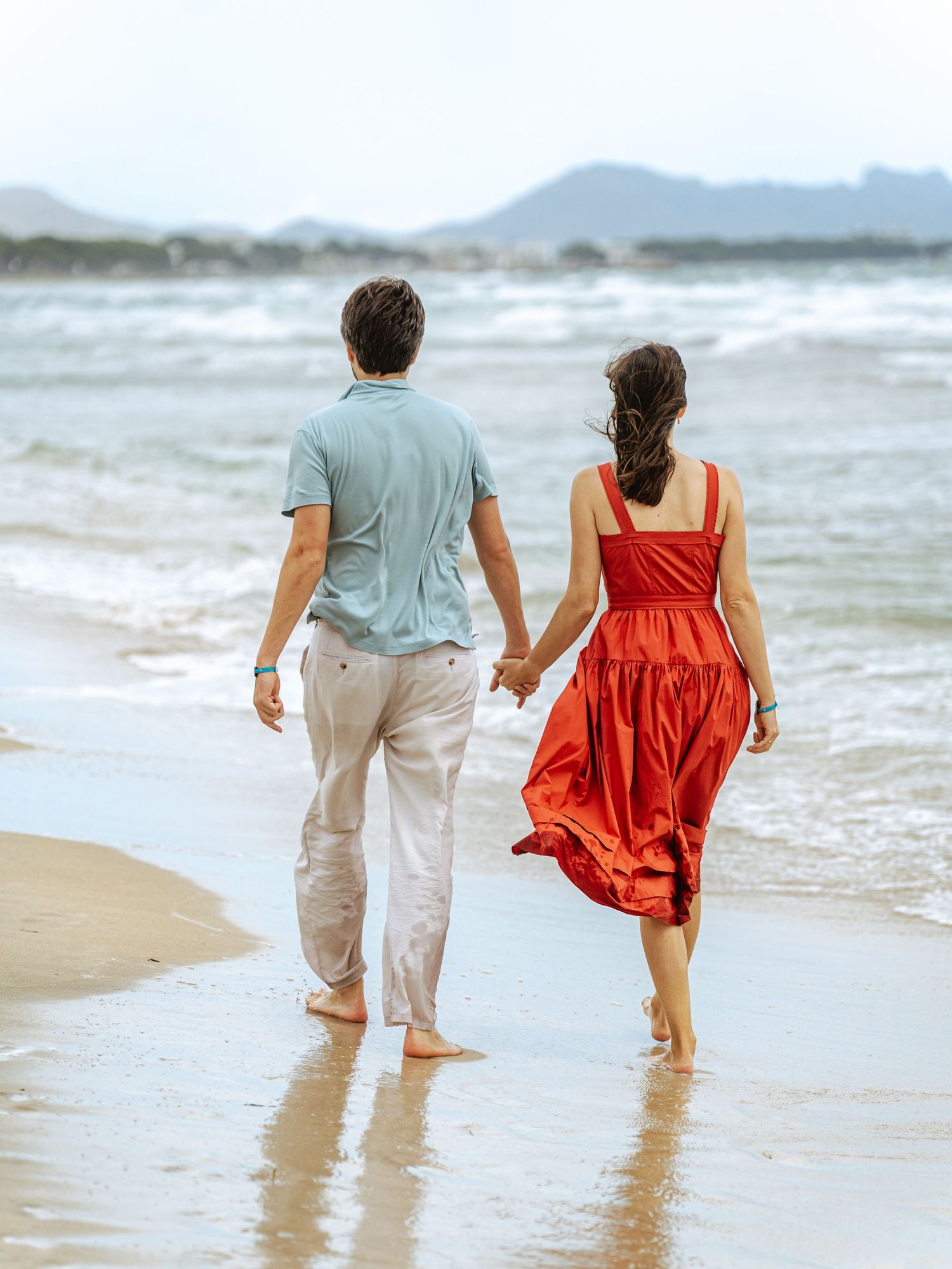 A couple holds hands walking along a scenic beach shoreline with mountains visible in the distance.
