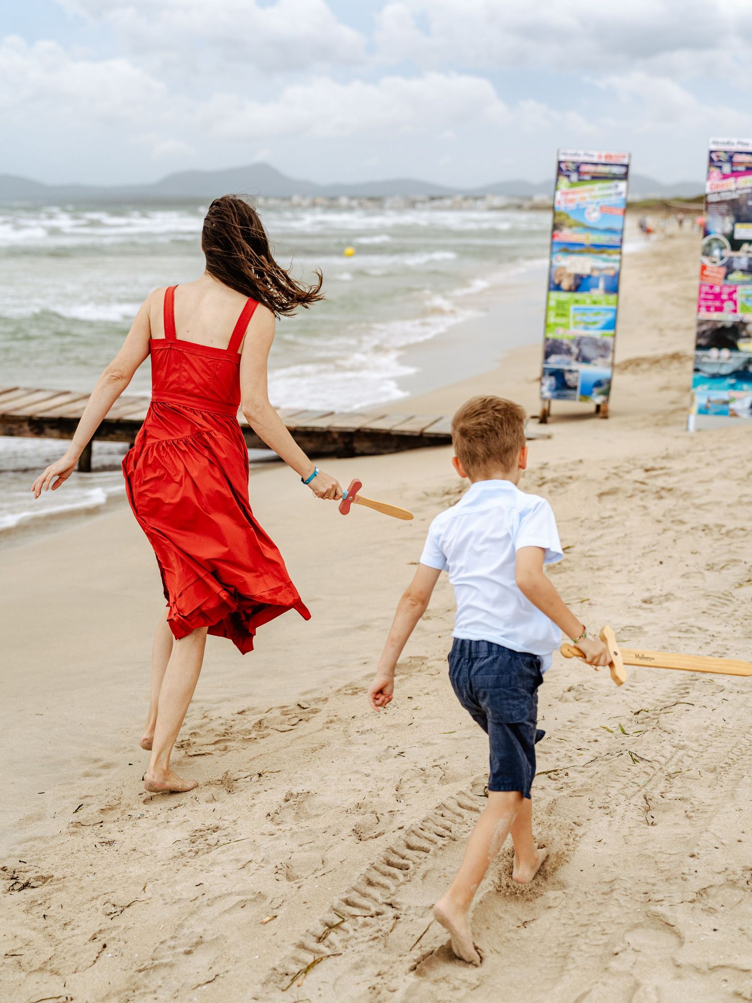 Woman in red dress and boy running on sandy beach toward ocean waves.