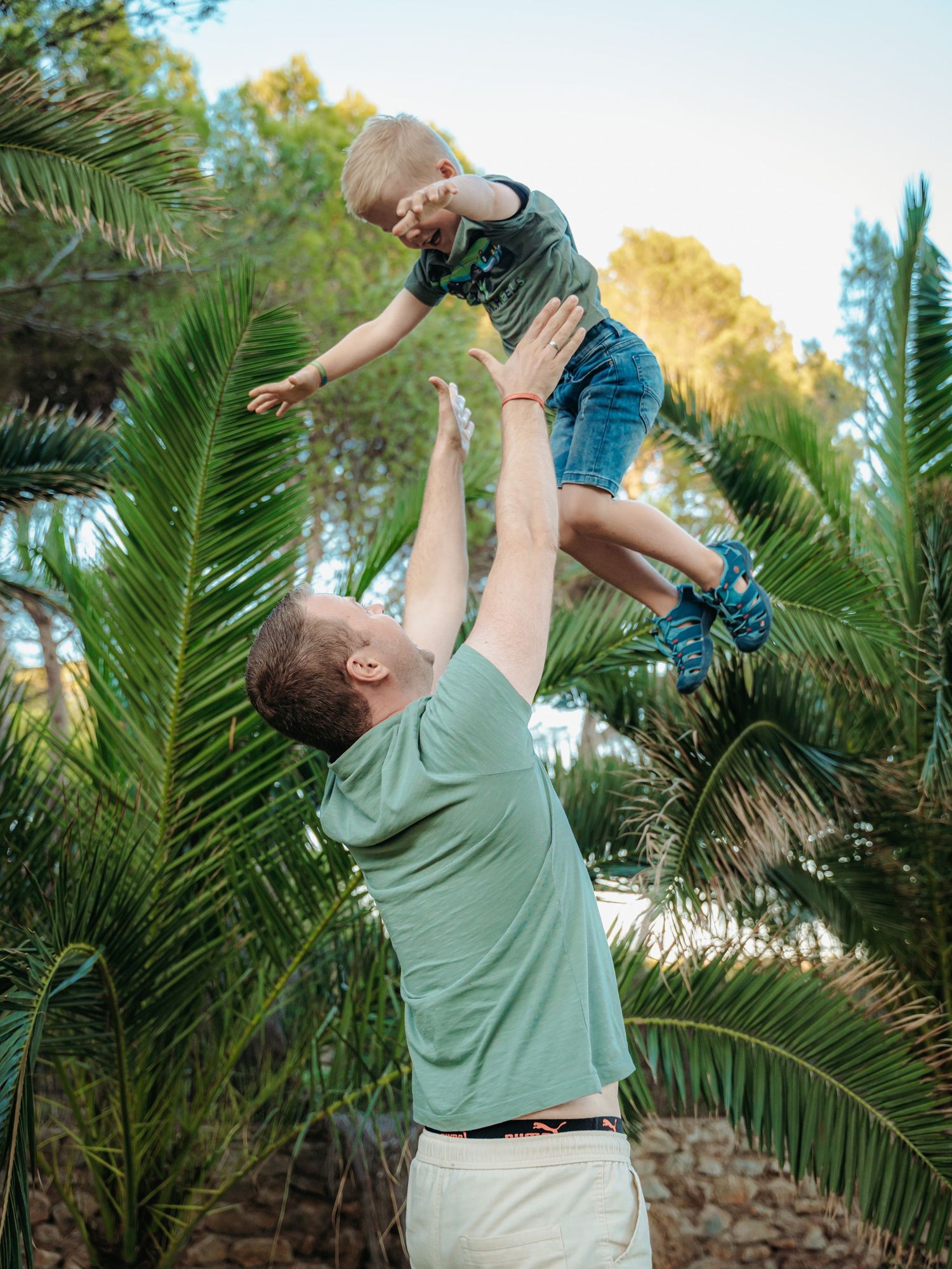 A father tosses his young son high in the air surrounded by lush green palm trees outdoors.