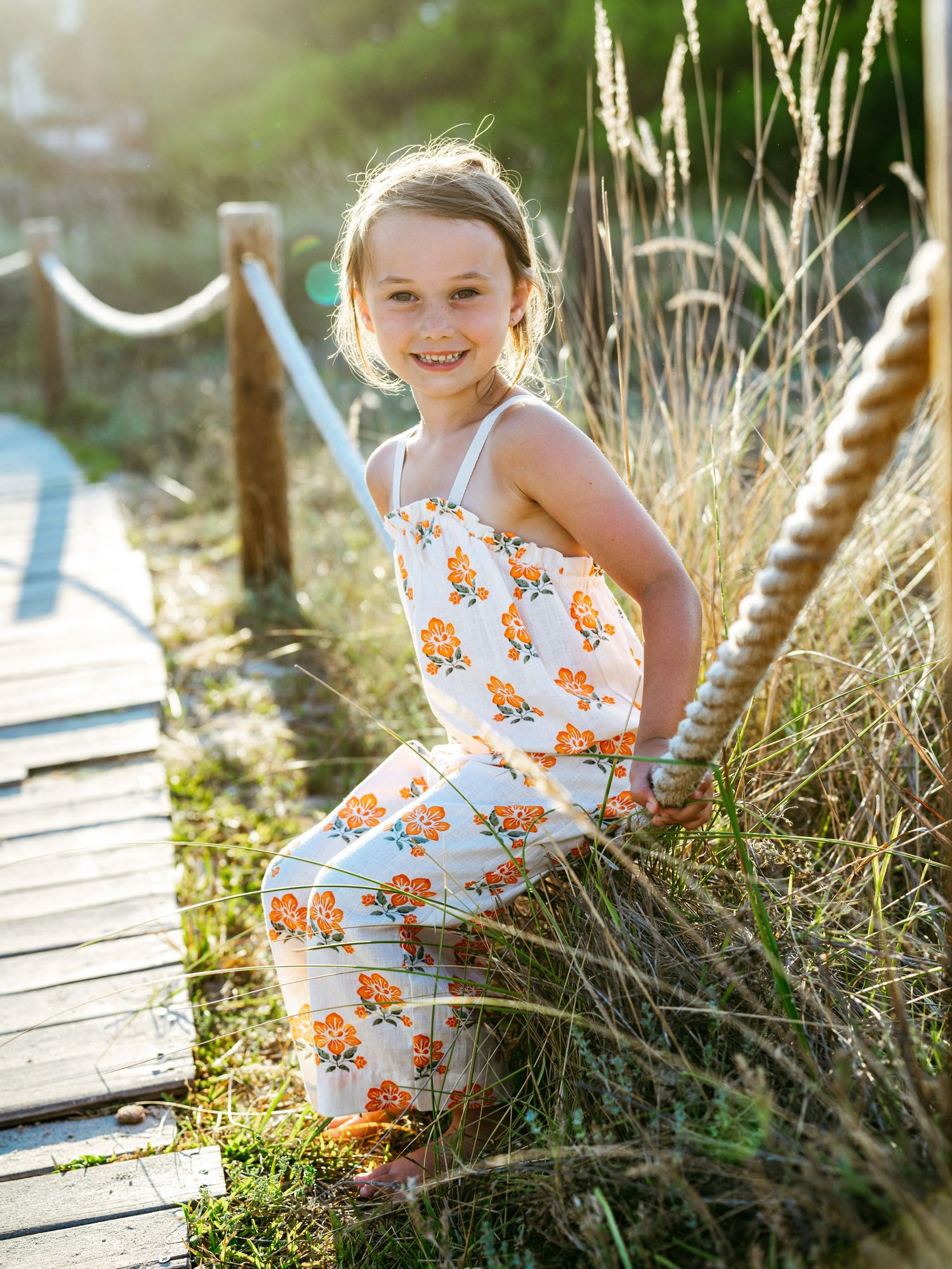 Smiling young girl in orange floral jumpsuit sitting on a sunny boardwalk surrounded by tall grass.