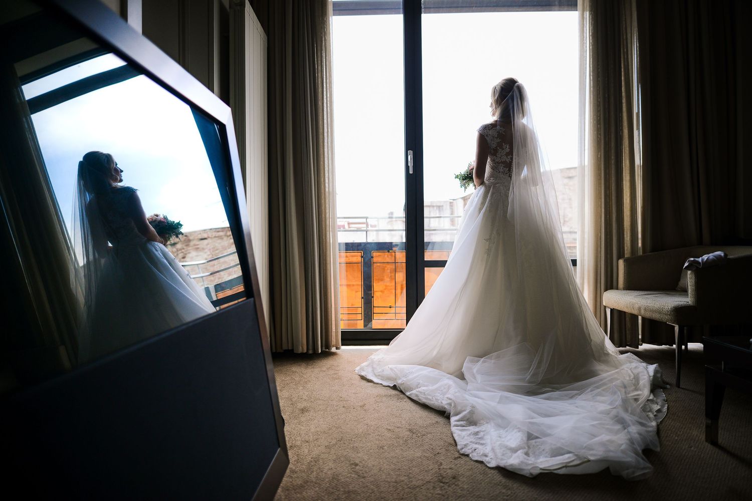 Bride standing by a hotel window before her wedding ceremony, reflected in a mirror, elegant bridal portrait, Herzenshaus Studio, Frankfurt.