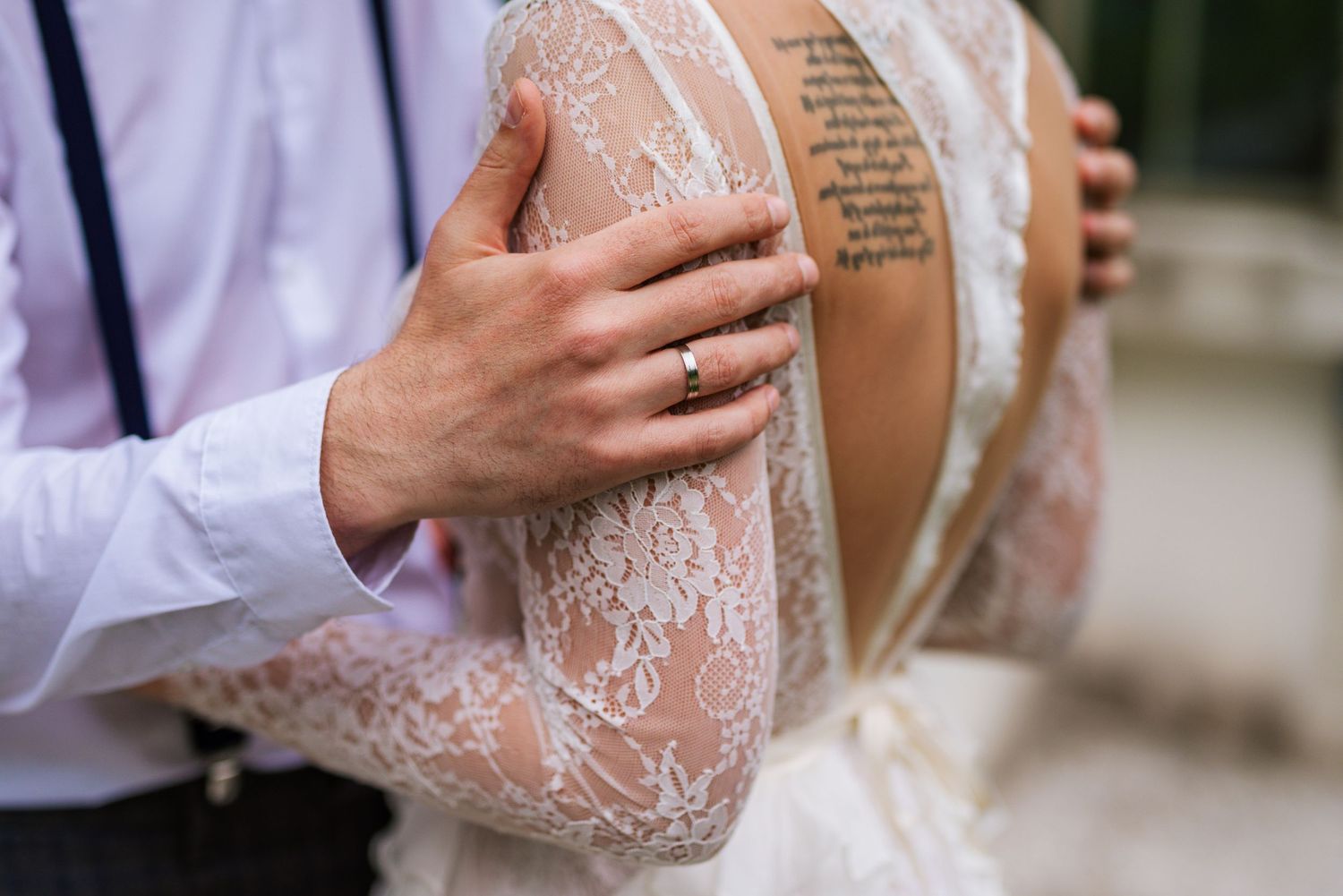 Close-up of groom's hand with wedding ring resting on bride's lace dress over her tattoo.