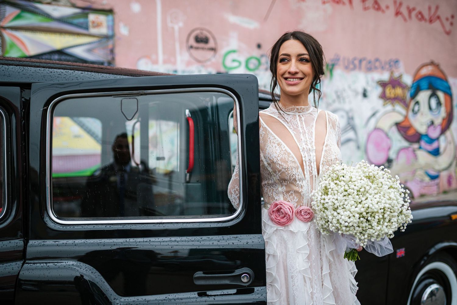 Radiant bride in lace gown holding white baby's breath bouquet beside black vintage car on wedding day.