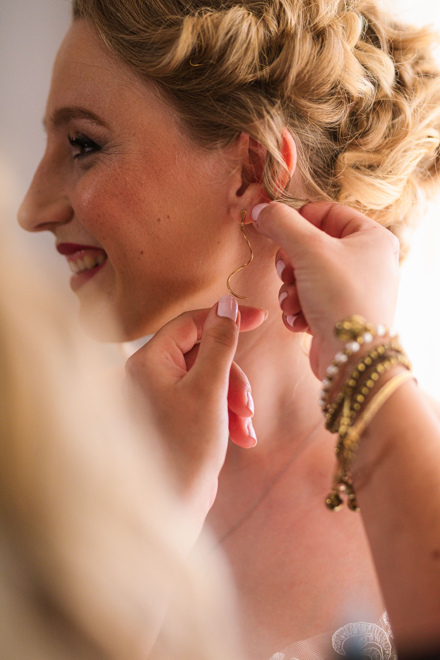 Close-up of bridesmaid carefully placing a gold earring on smiling bride's ear.