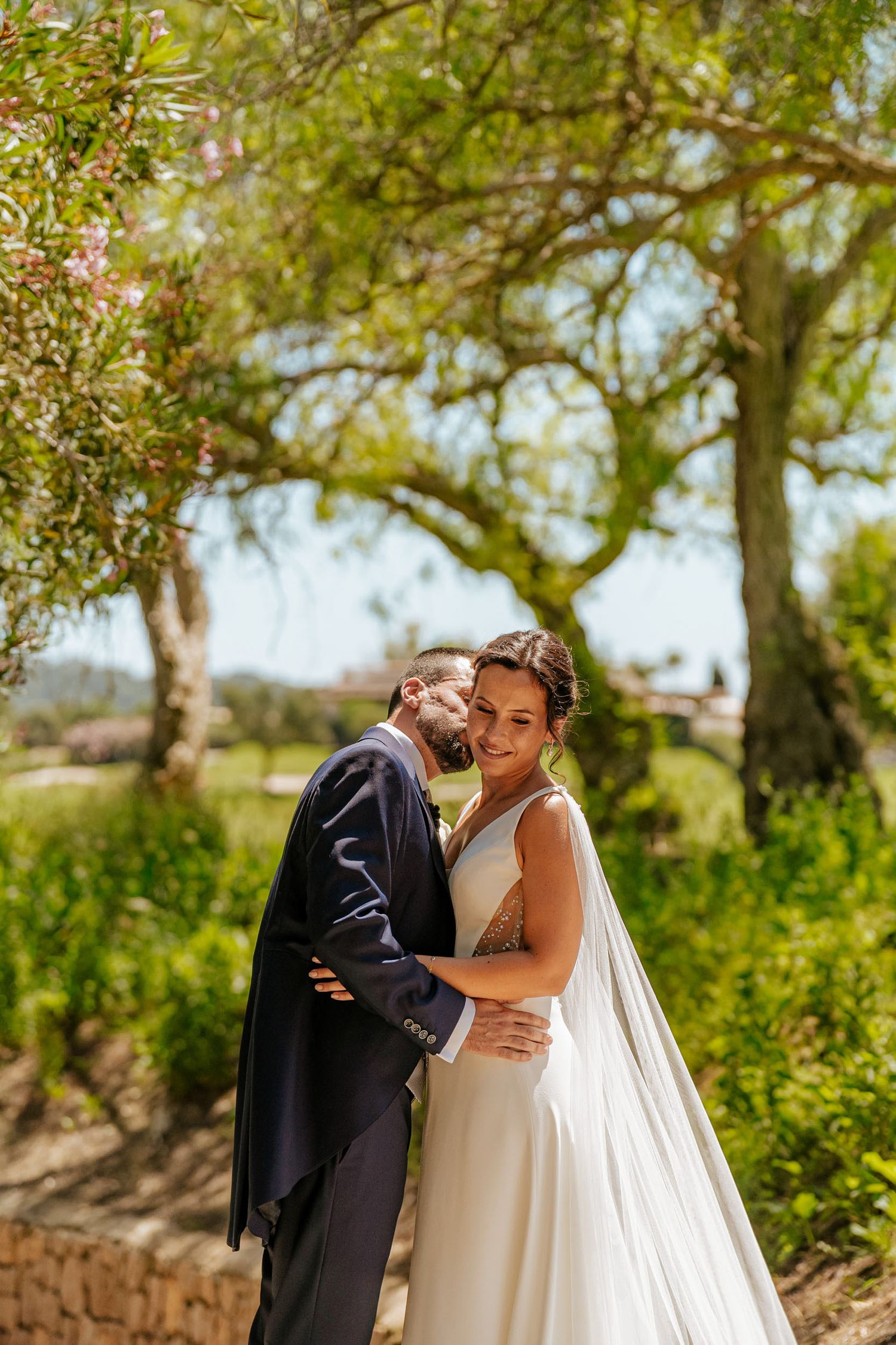 Groom tenderly embraces smiling bride among lush green trees in sunlit outdoor setting.