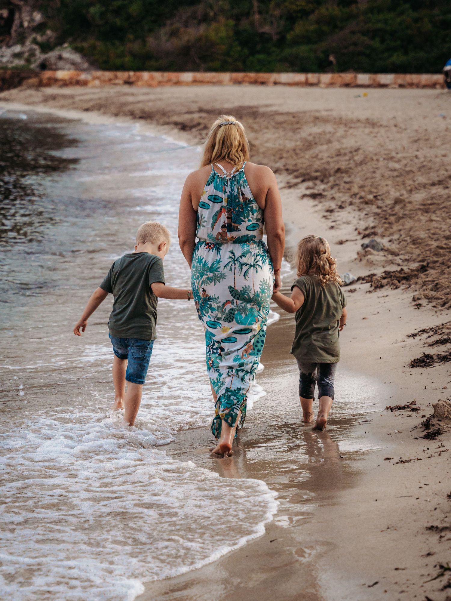 Mother in tropical floral dress walking hand in hand with two young children along ocean shoreline.