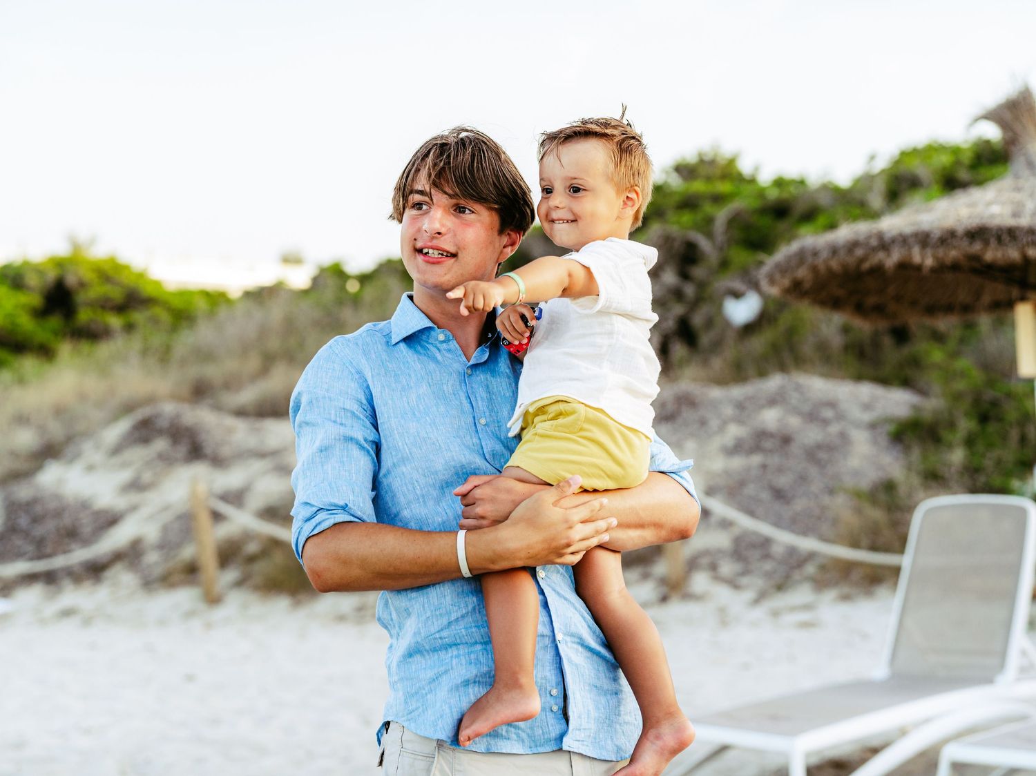 Young man in blue linen shirt holding a smiling toddler on a sandy beach with dunes behind them.