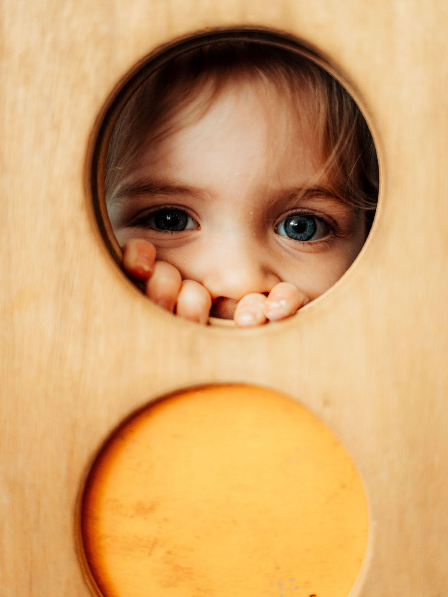 A curious young child peers through a circular hole in a wooden play structure, eyes wide with wonder.