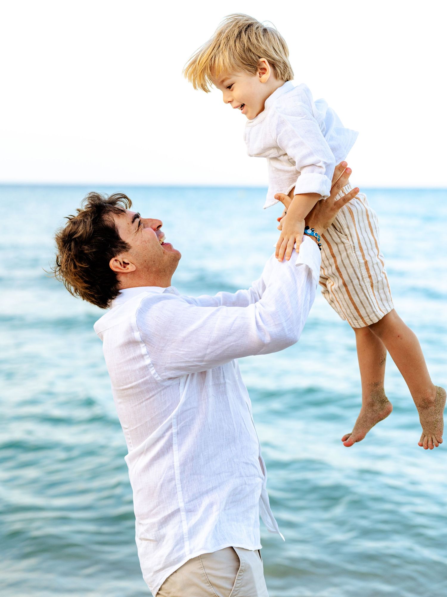 Father in white linen shirt lifting happy blond child at seaside.