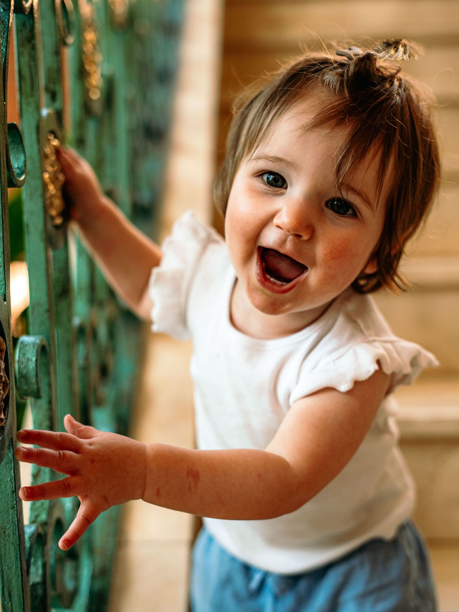 Happy toddler girl laughing while holding green ornate iron fence outdoors in natural light.