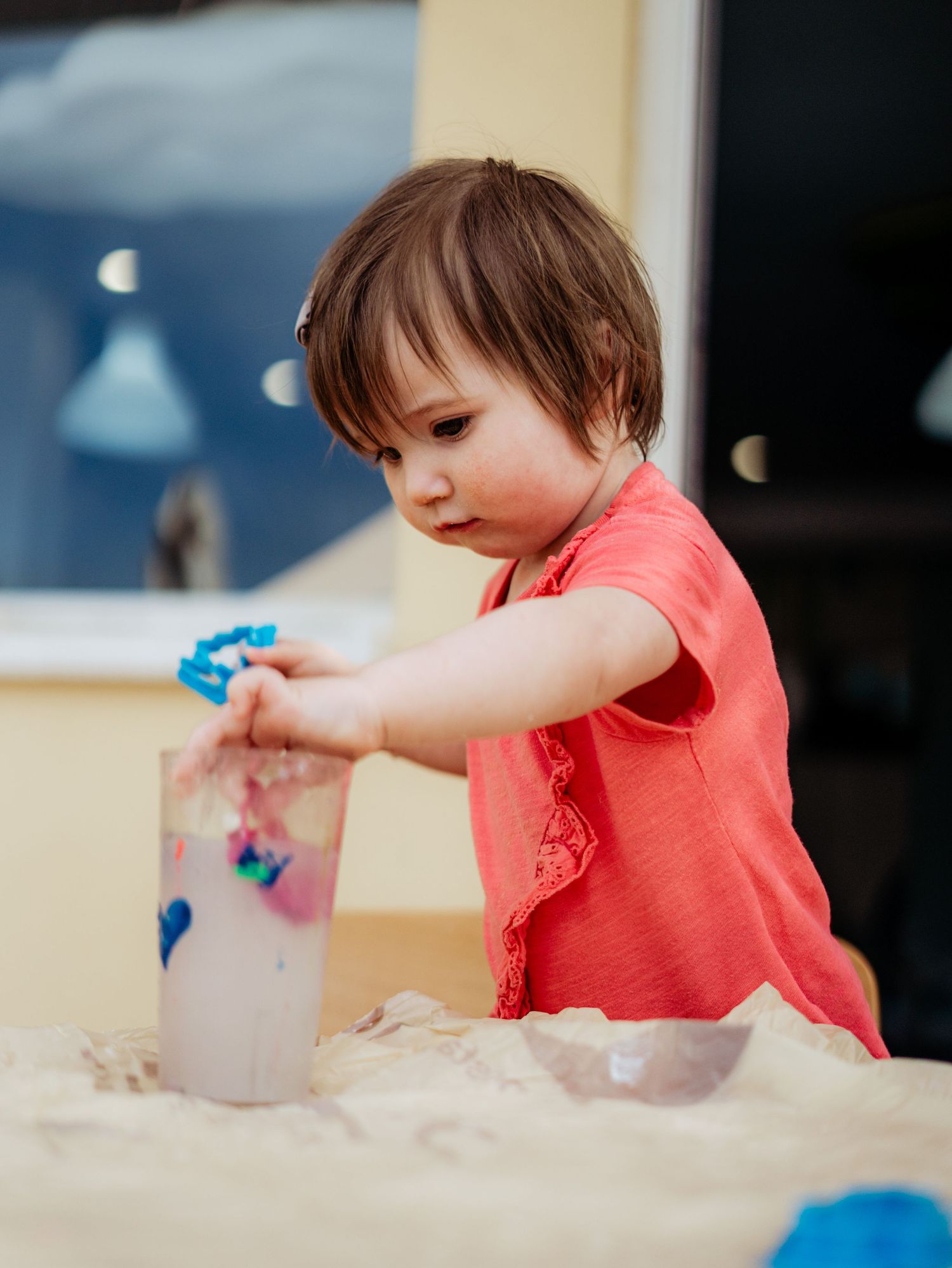 Young toddler girl in coral shirt focused on placing items into a decorated cup at a table.