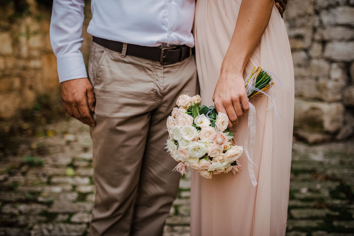 Close-up of bride in blush gown holding elegant white and pink floral bouquet beside groom.