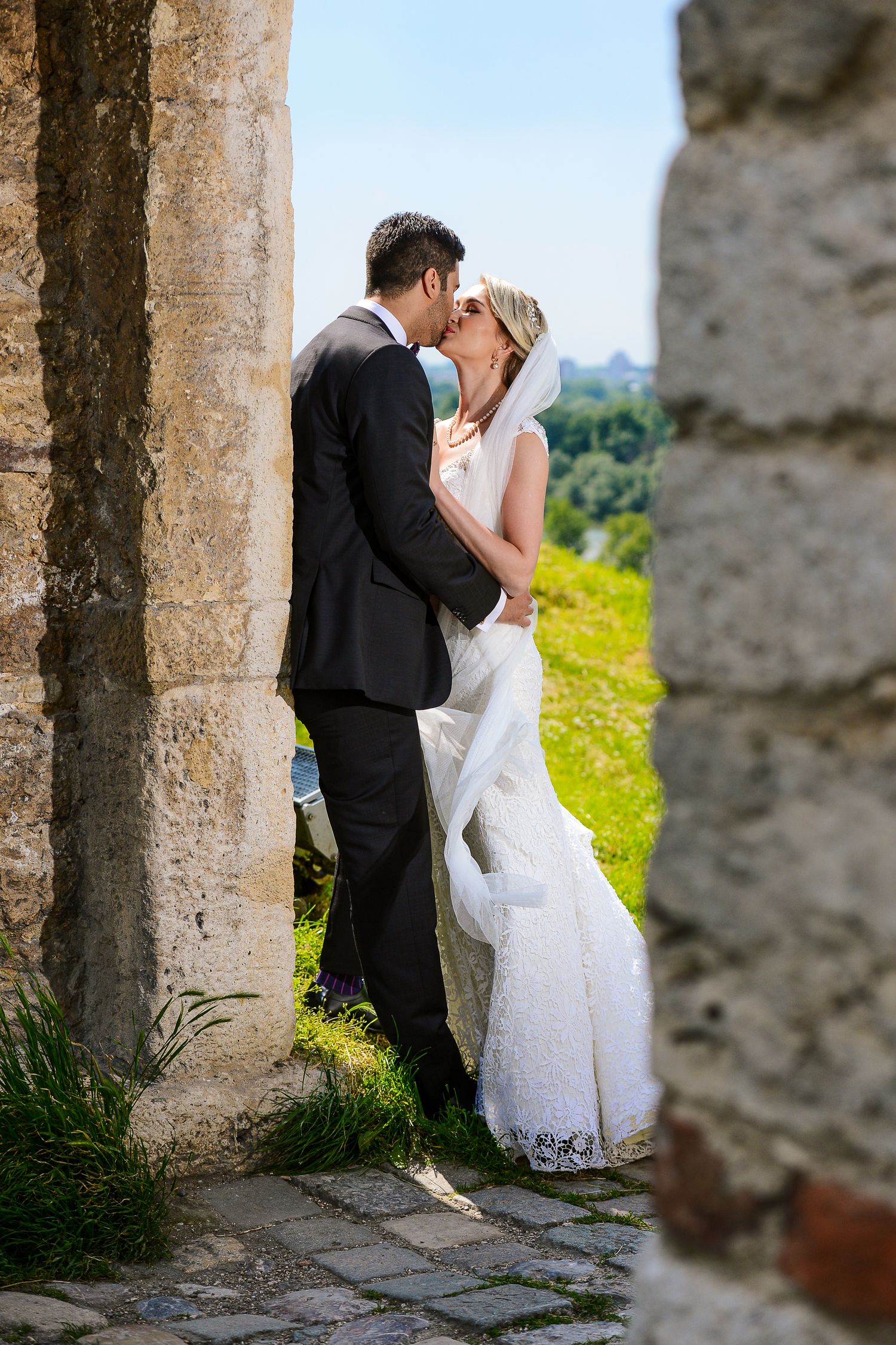 Bride and groom sharing a romantic kiss between ancient stone columns outdoors on their wedding day.