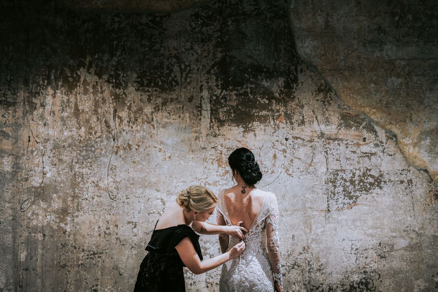 Woman in black dress fastening buttons on bride's backless lace wedding gown outdoors.
