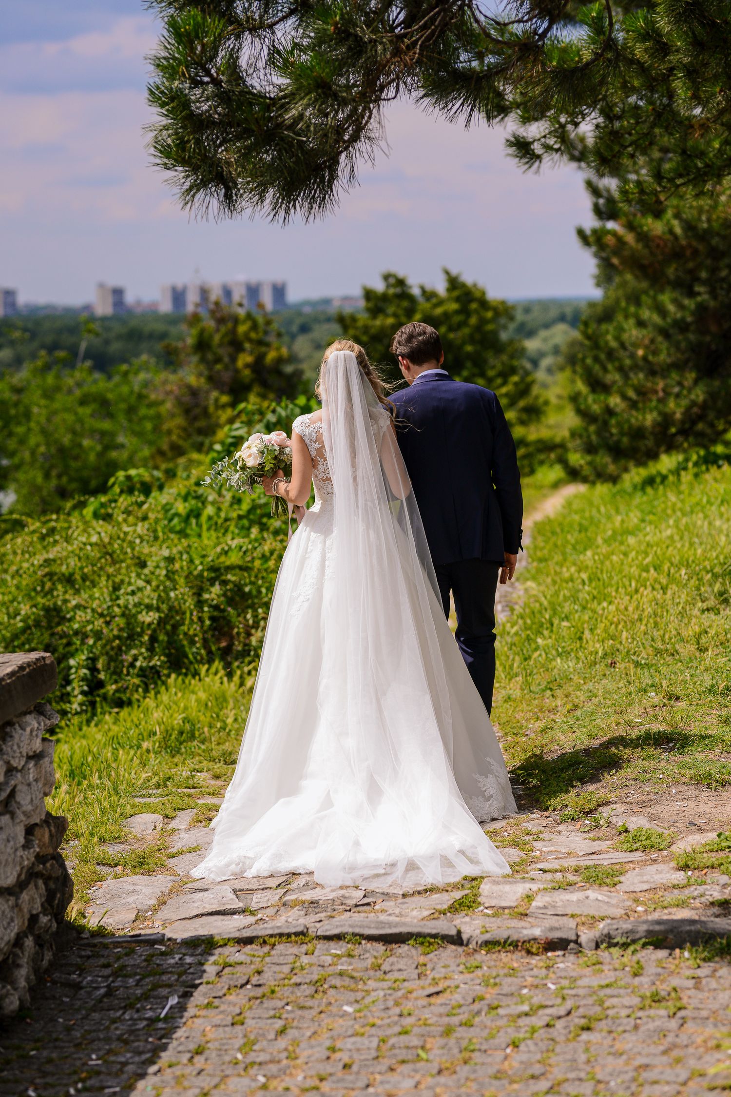 Bride and groom walk hand in hand along a cobblestone path with a scenic green landscape behind them.