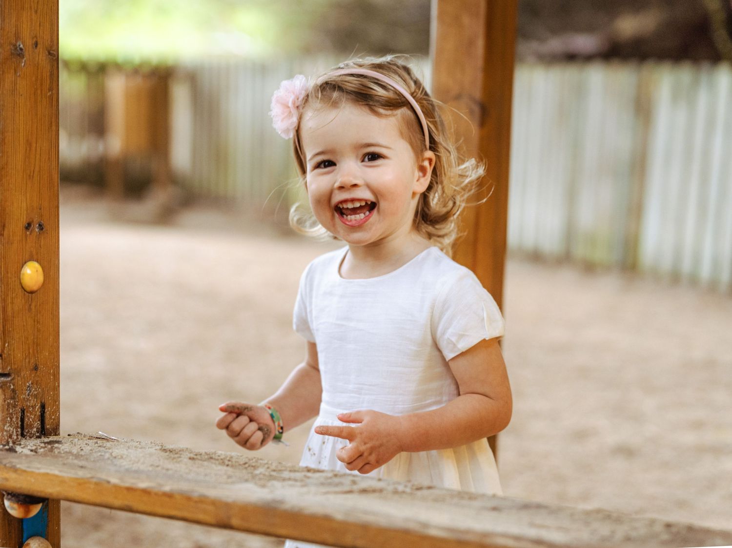 Joyful toddler girl in white dress laughing at outdoor wooden playground in sandy area.