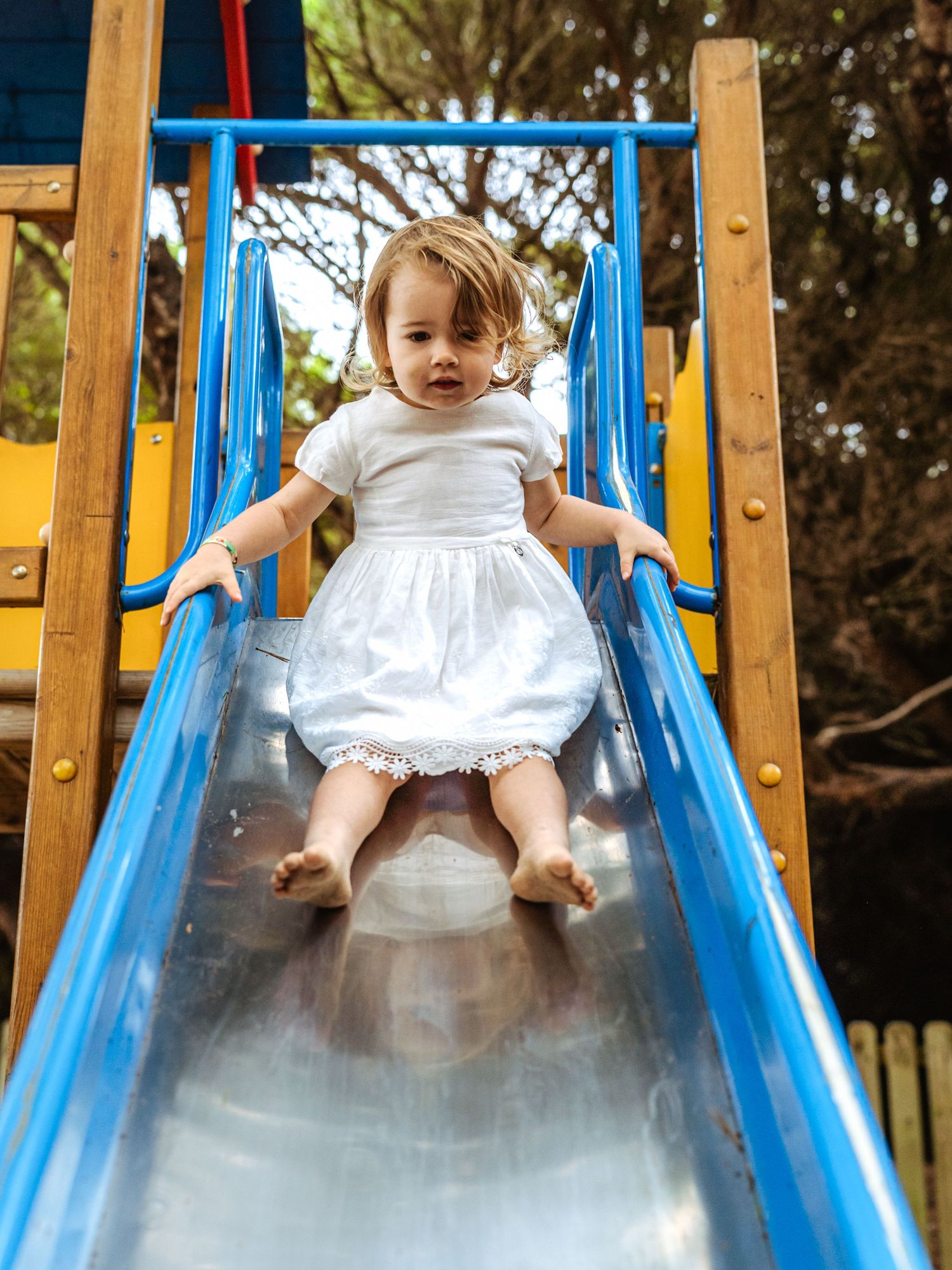 A cute toddler girl in a white dress slides down a blue metal playground slide barefoot.