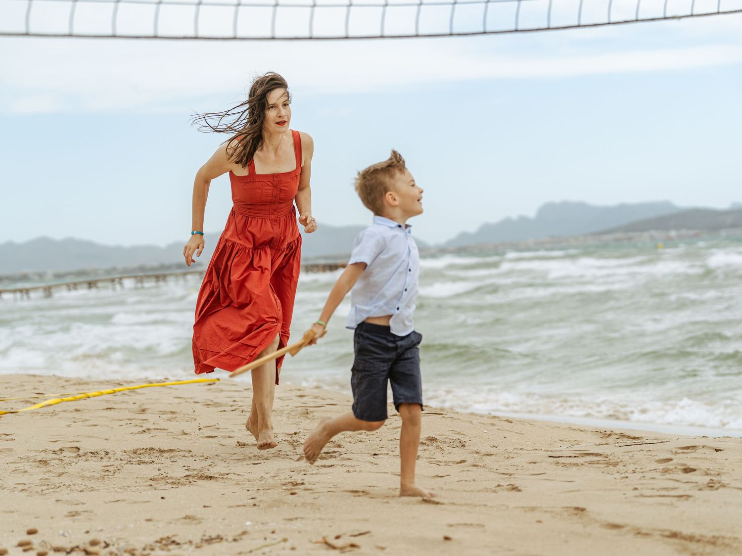 Mother in red dress running with young boy along sandy beach near volleyball net.