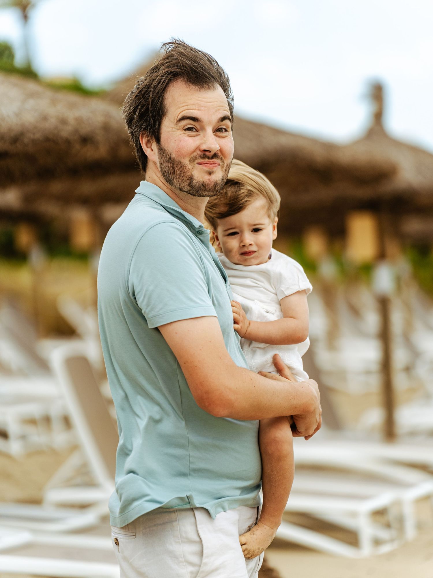 Smiling father holding young toddler near beach lounge chairs and thatched umbrellas.