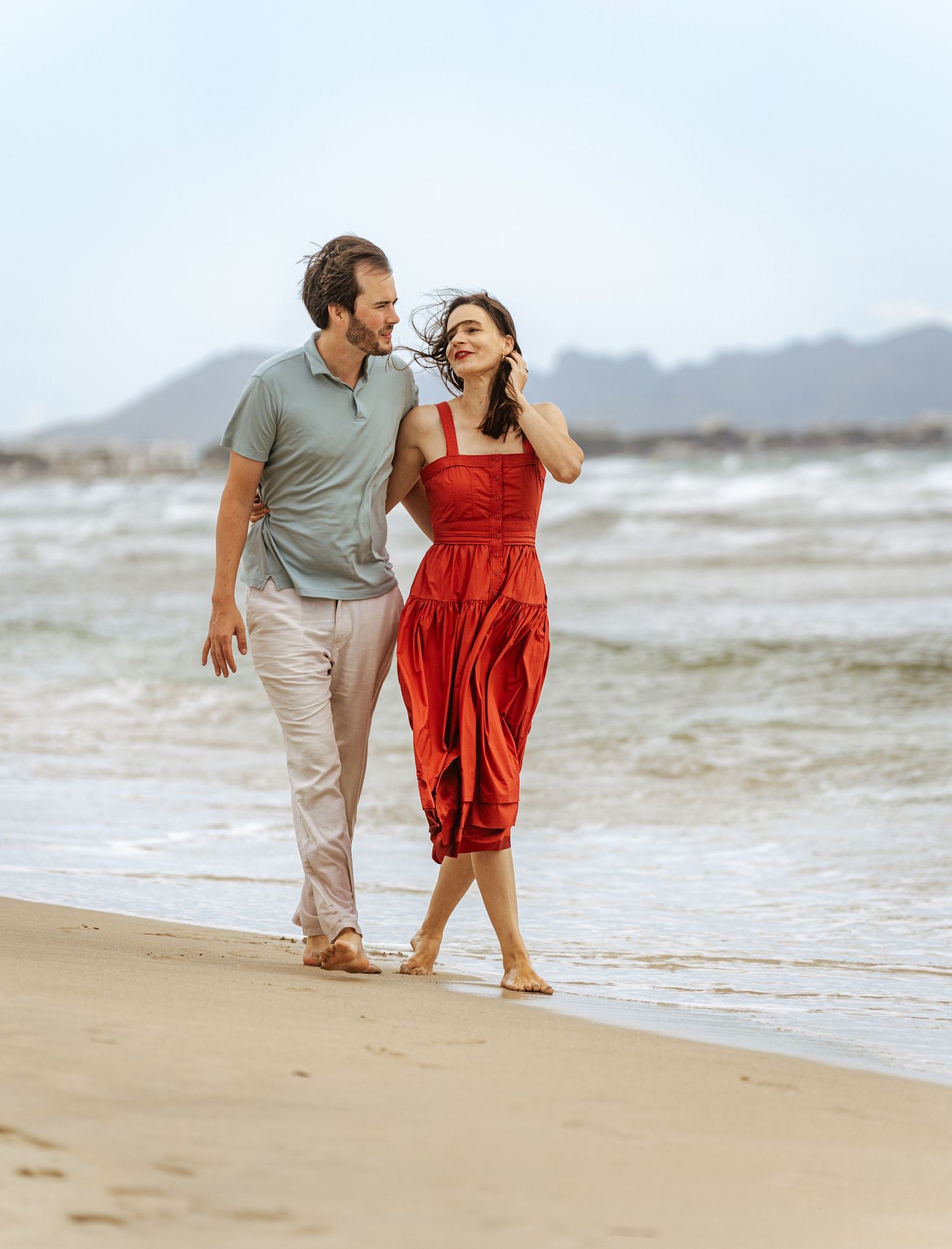 Joyful couple strolling along a windy beach shoreline, woman in flowing red dress, laughing together.