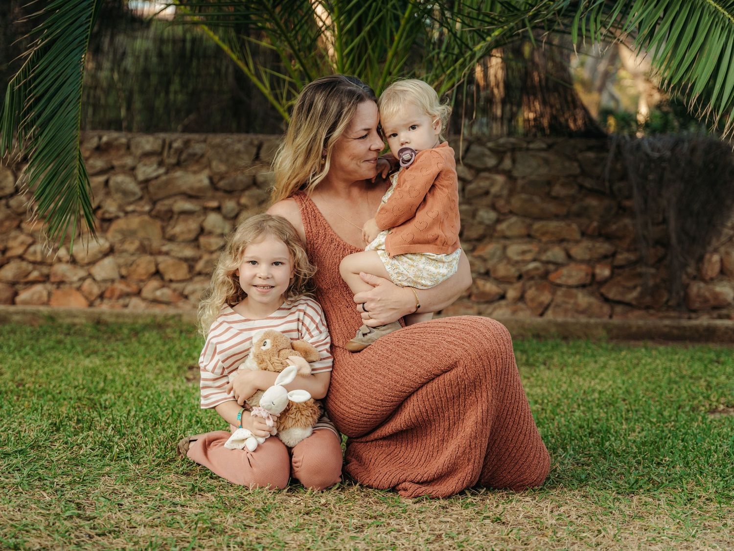 Mother sitting on grass kissing baby while young girl holds teddy bear near palm tree.